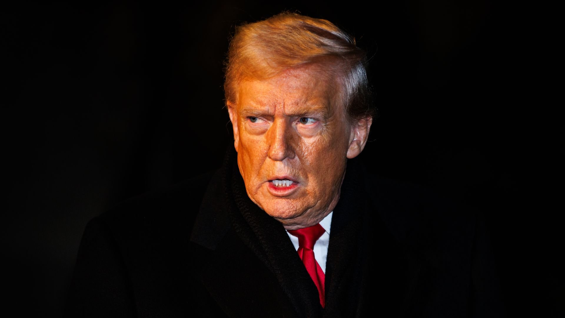 A close-up of a man with orange-toned skin and blond hair, wearing a black coat, red tie, and white shirt, looking to the side with a serious expression against a dark background.