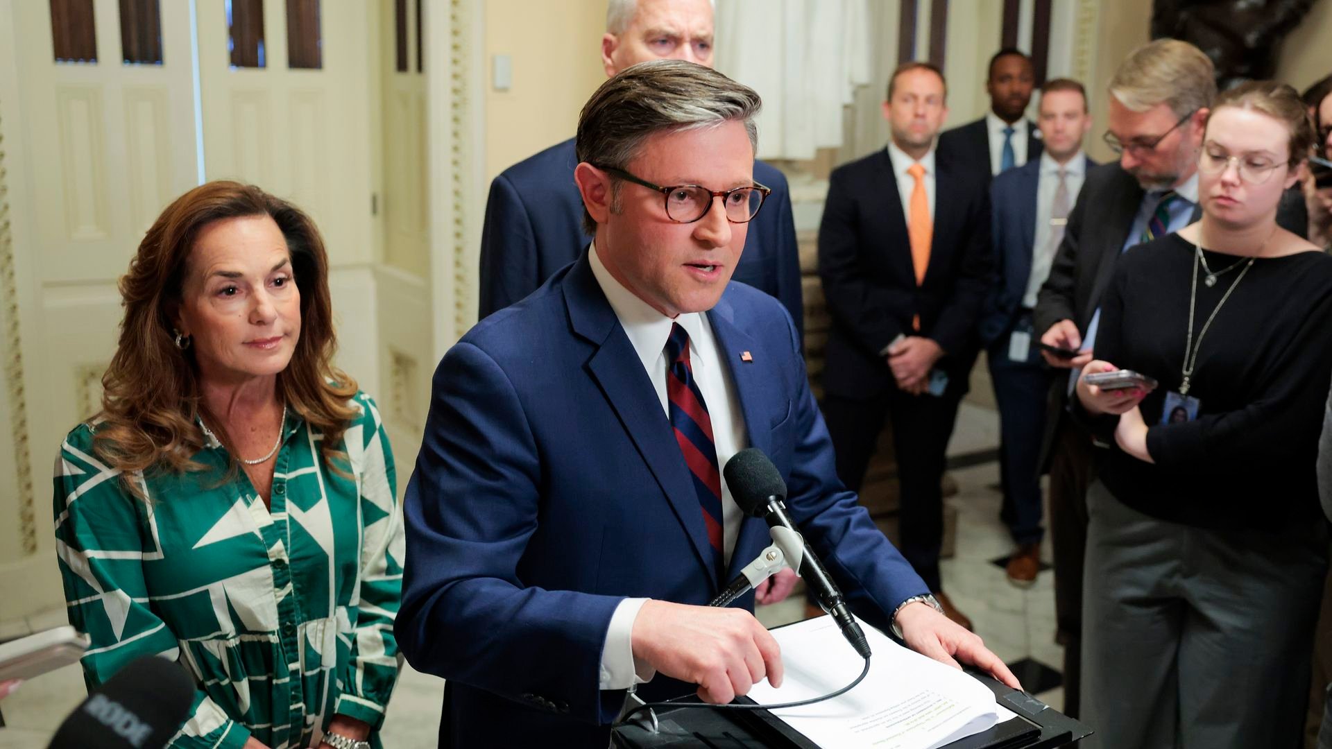 Man in a blue suit speaks at a podium with a microphone and papers, flanked by a woman in a green patterned dress; reporters observe in the background.