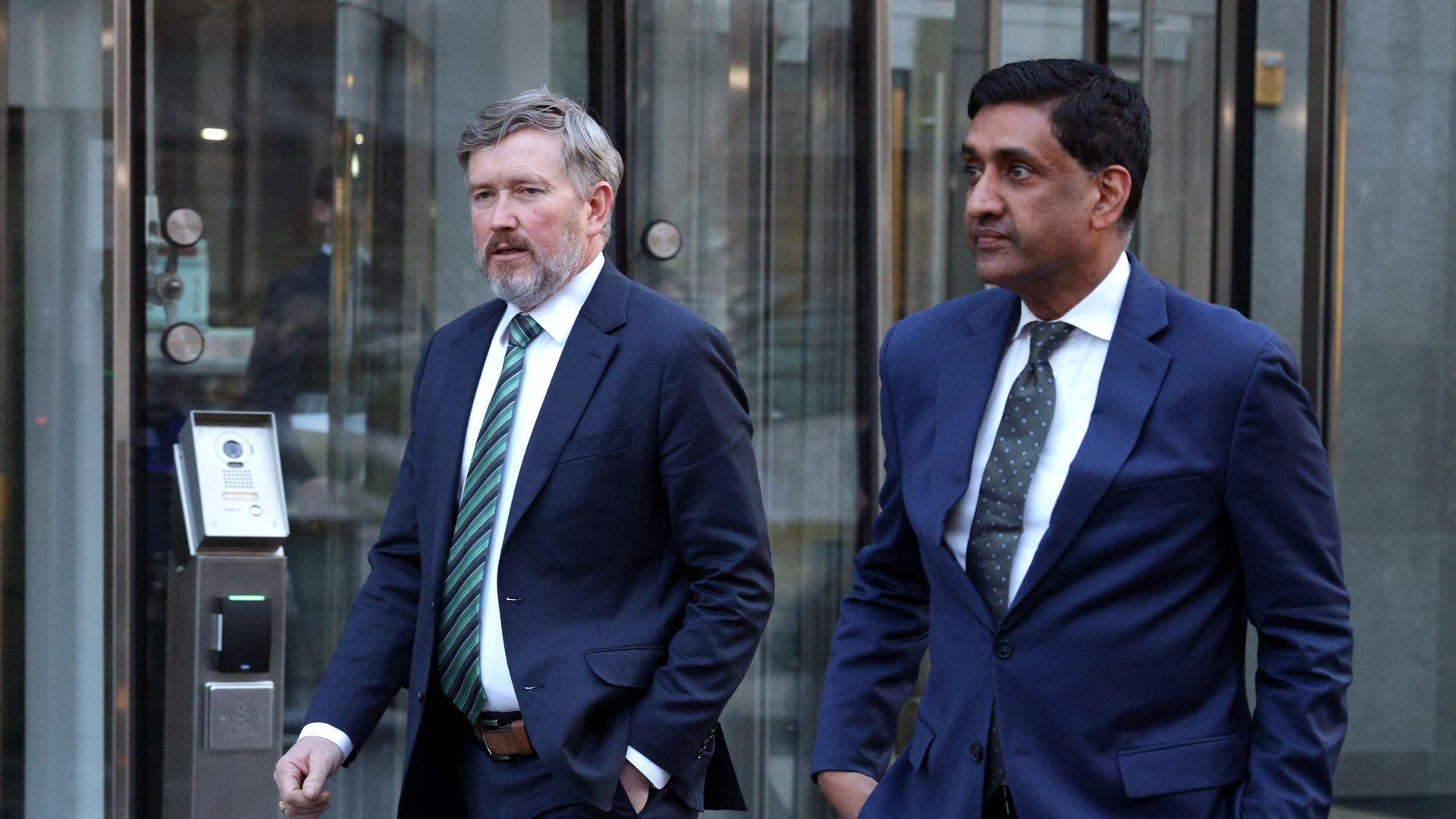 Two men in navy suits and ties, one gray-haired with a striped tie, the other dark-haired with a dotted tie, walking outside a glass building entrance.