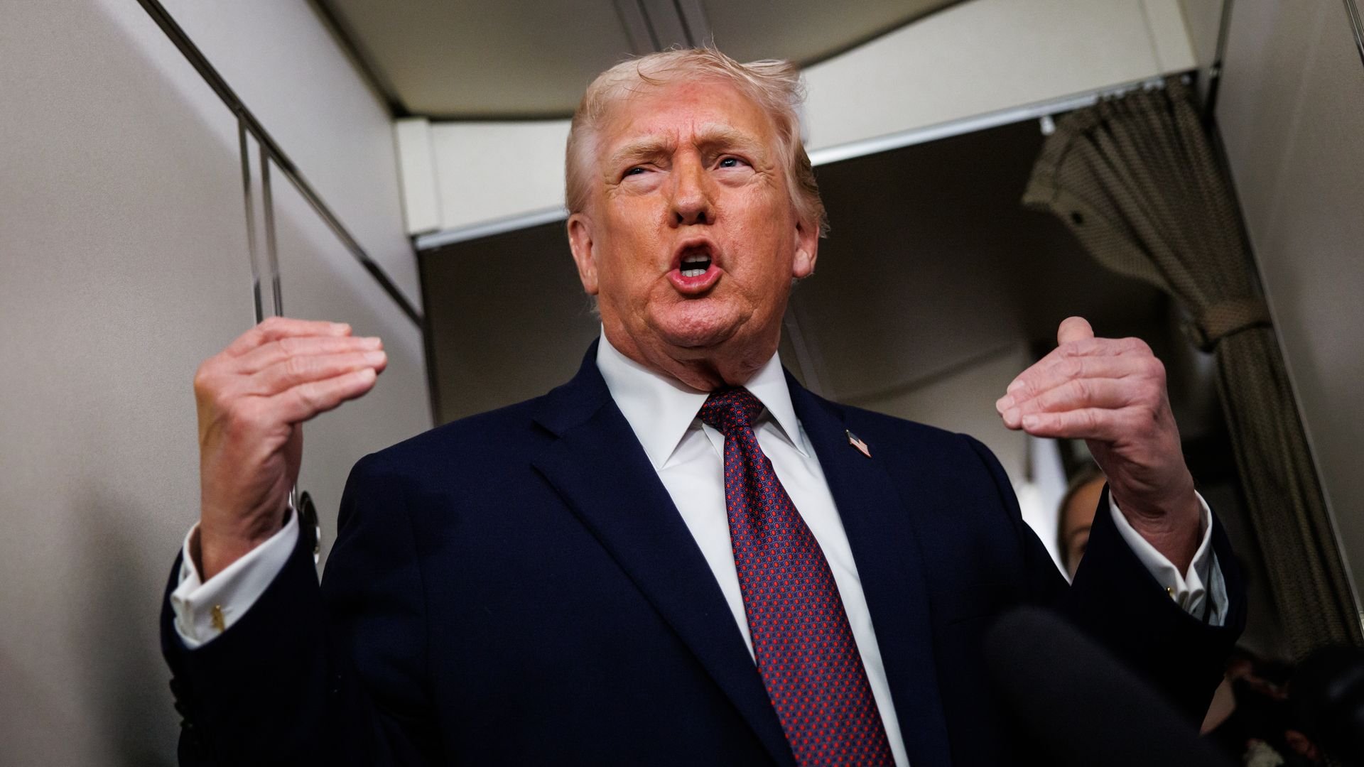 Man in dark blue suit and red patterned tie speaking with hands raised inside an airplane cabin