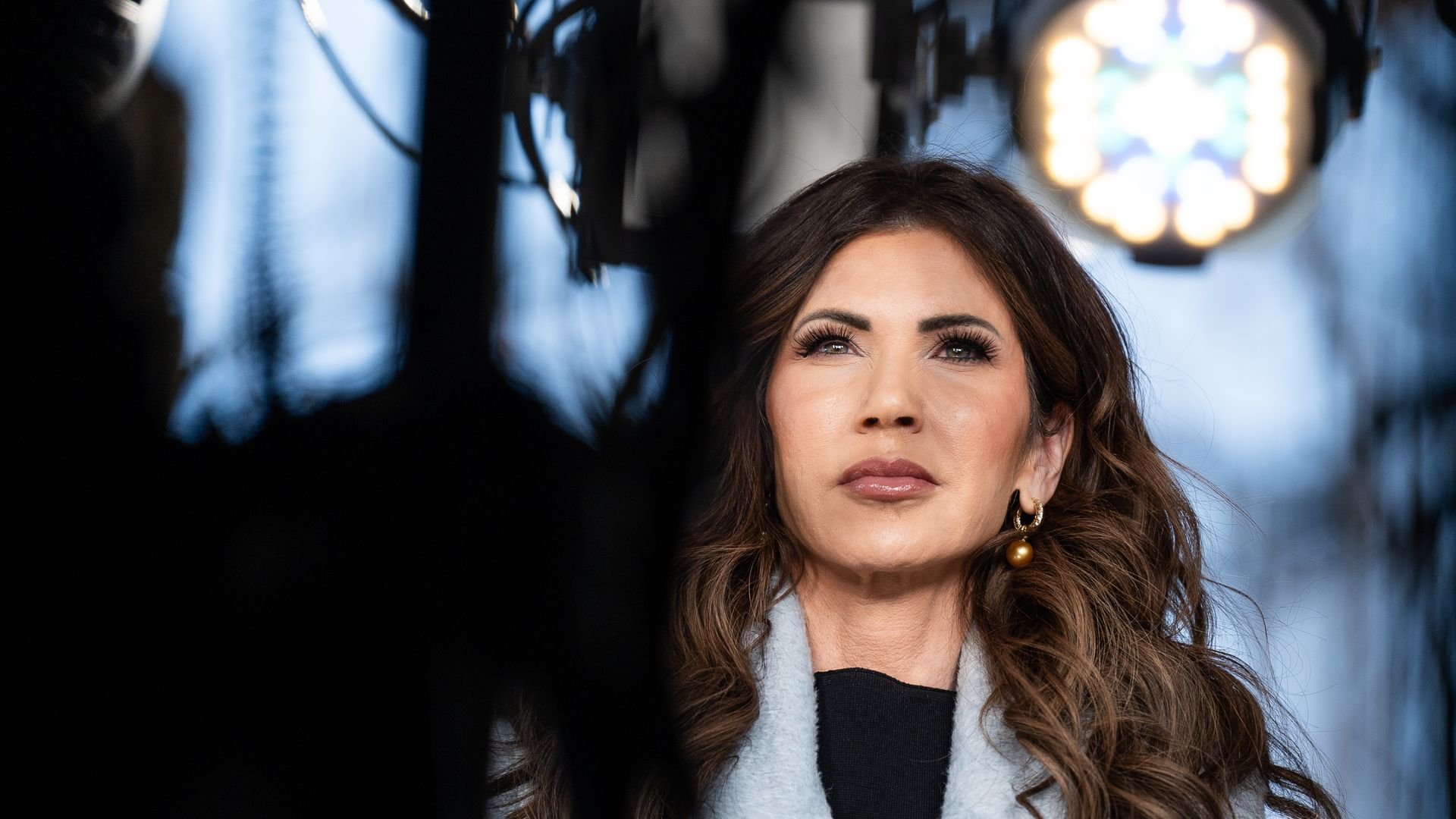 Close-up of a woman with long brown hair and gold earrings, wearing a light gray coat over black, standing under stage lighting with a blurred blue background.