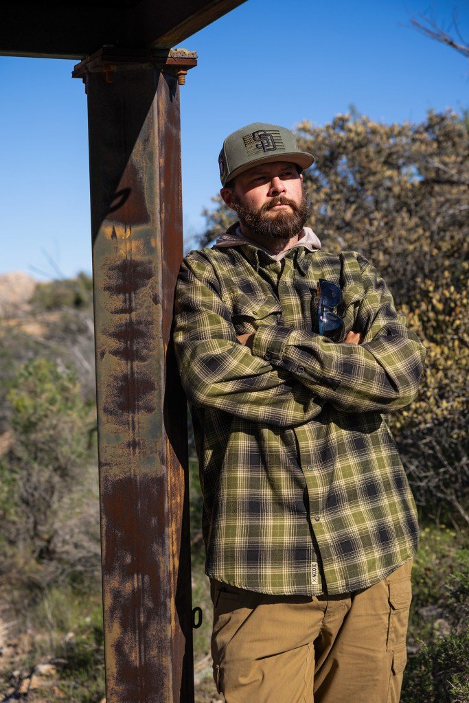 A person wearing a baseball cap and plaid shirt stands with arms crossed beside a rusted metal post outdoors, with shrubs and a clear blue sky in the background.