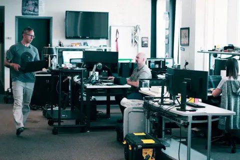 Greg Squire and colleagues including Pete Manning in their office. Greg is wearing a green t-shirt and light trousers and is walking across a room crowded with computer screens, holding a laptop. 