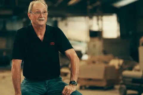 John Harp has short grey hair and a grey moustache. He is wearing a black short-sleeved shirt and green trousers, and is sitting in his brick factory, open boxes open behind him. 