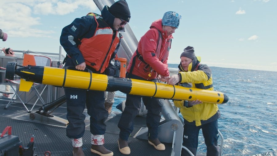 Ella Wawrzynek, Madeline Miller, and David Whelihan in winter clothing on a ship prepare to lower a long underwater vehicle into the ocean.