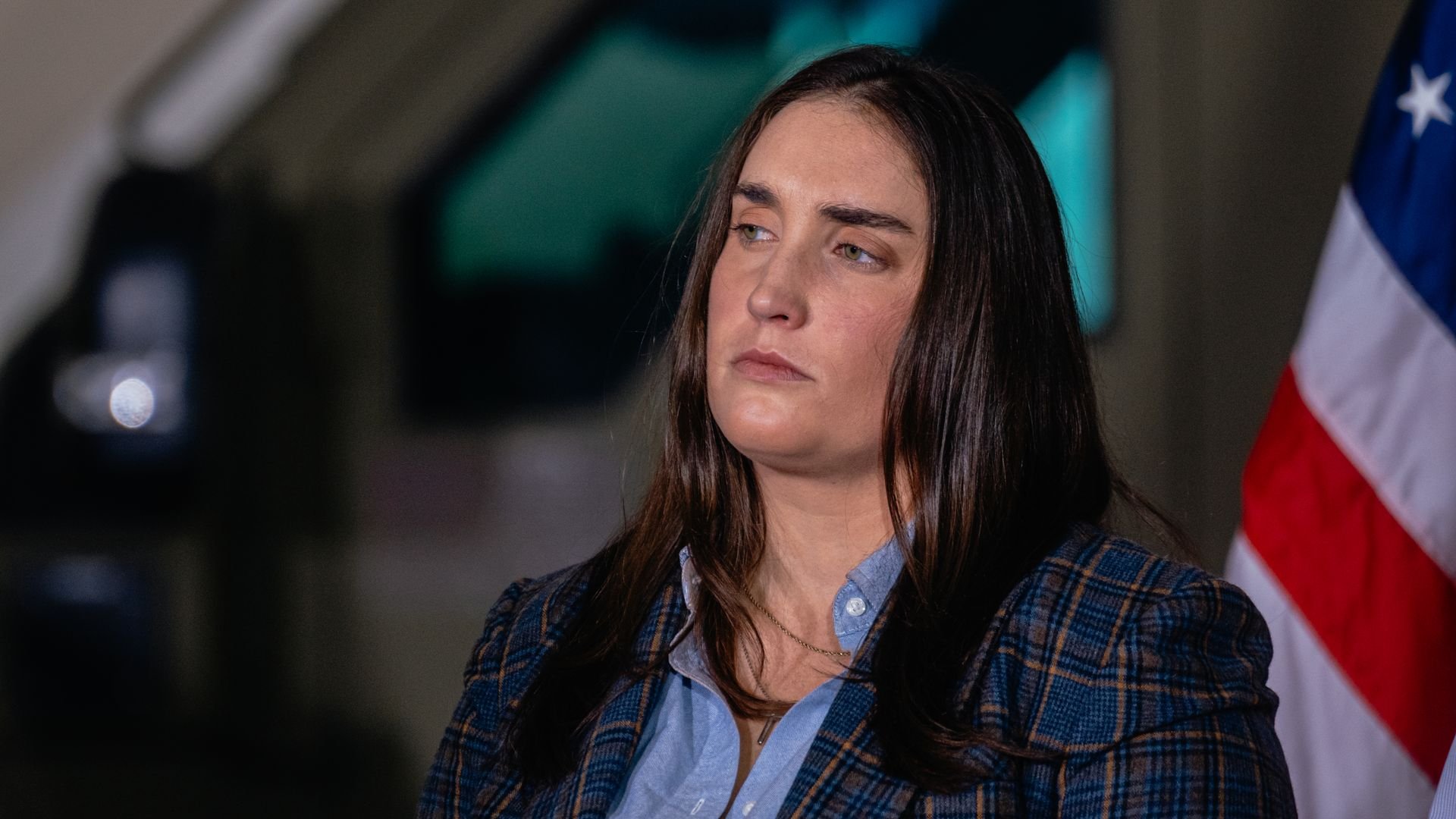 Woman with long brown hair wearing a blue shirt and blue plaid jacket, standing pensively near an American flag with a blurred background.