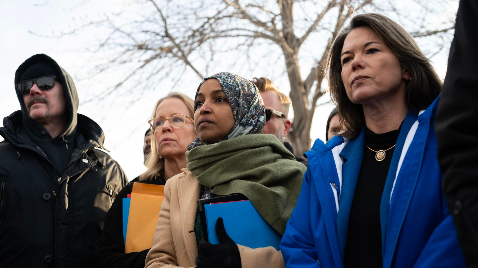 Reps. Ilhan Omar, Kelly Morrison and Angie Craig arrive outside the Bishop Henry Whipple Federal Building. The three women stand shoulder to shoulder, two of whom are holding folders, in a crowd of people outside an ICE facility.