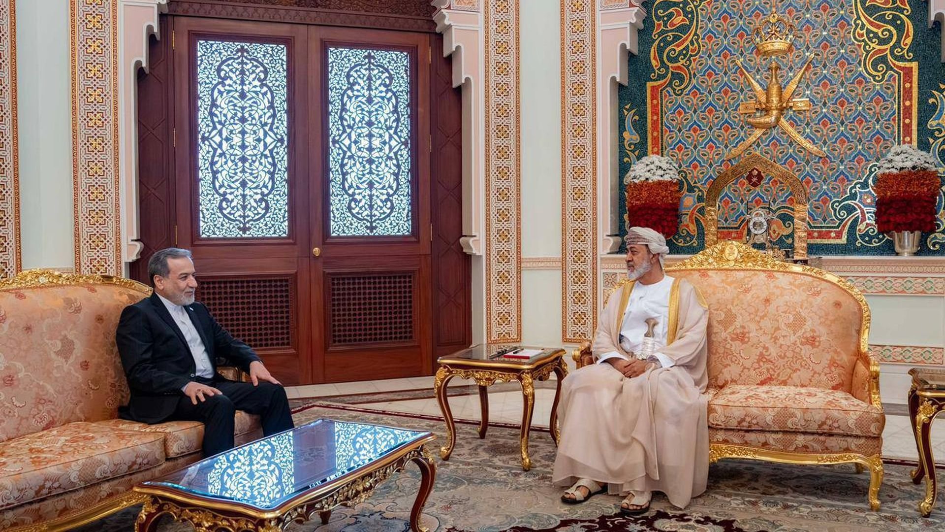Two men sit on ornate gold sofas in a luxurious room with intricate wallpaper, a glass coffee table, and a decorative wall behind. Left man wears a dark suit; right man wears white traditional robes.