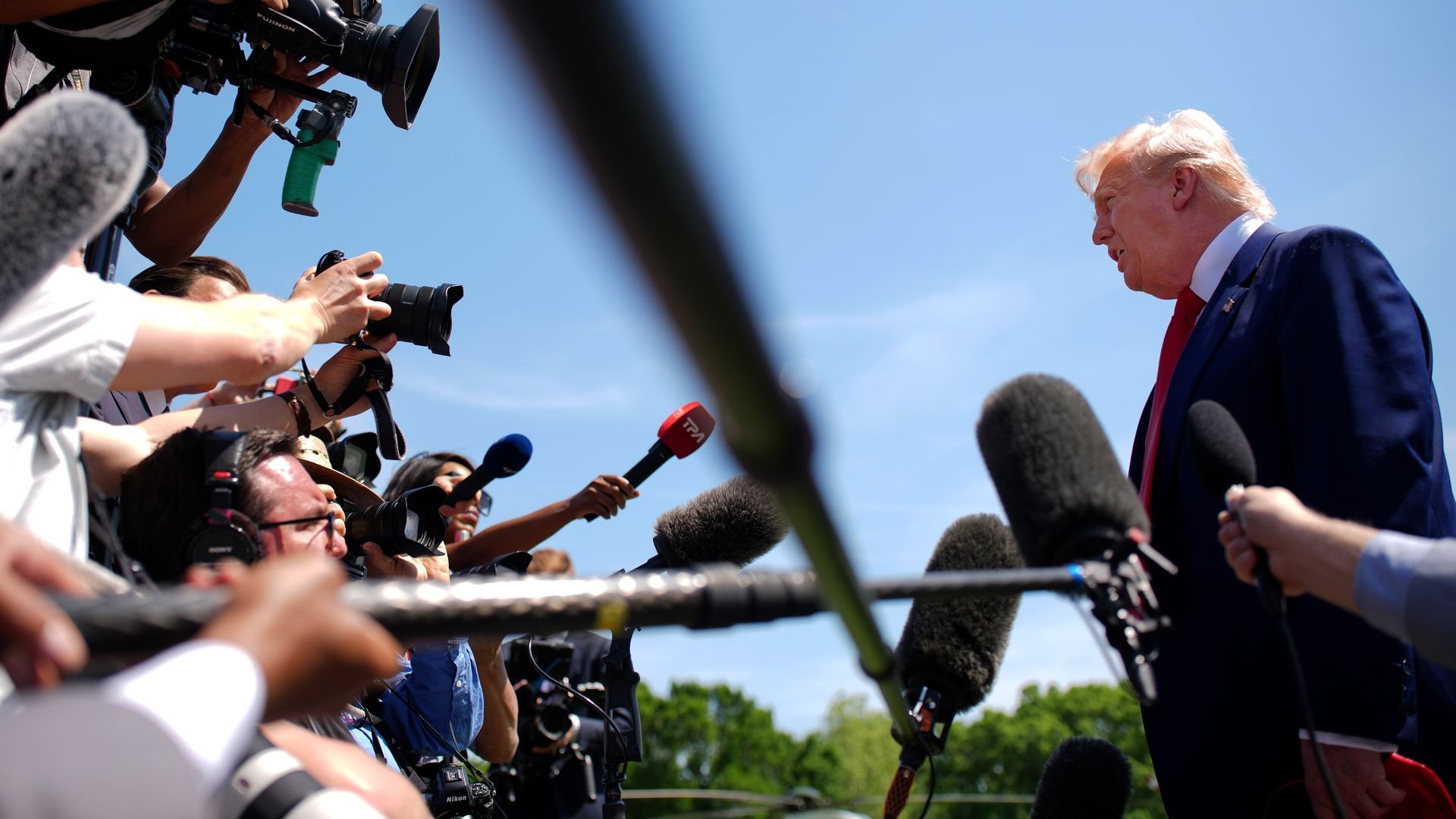 President Donald Trump stands outdoors on the White House South Lawn speaking to a crowd of reporters and cameras, with microphones extended toward him under a clear blue sky.