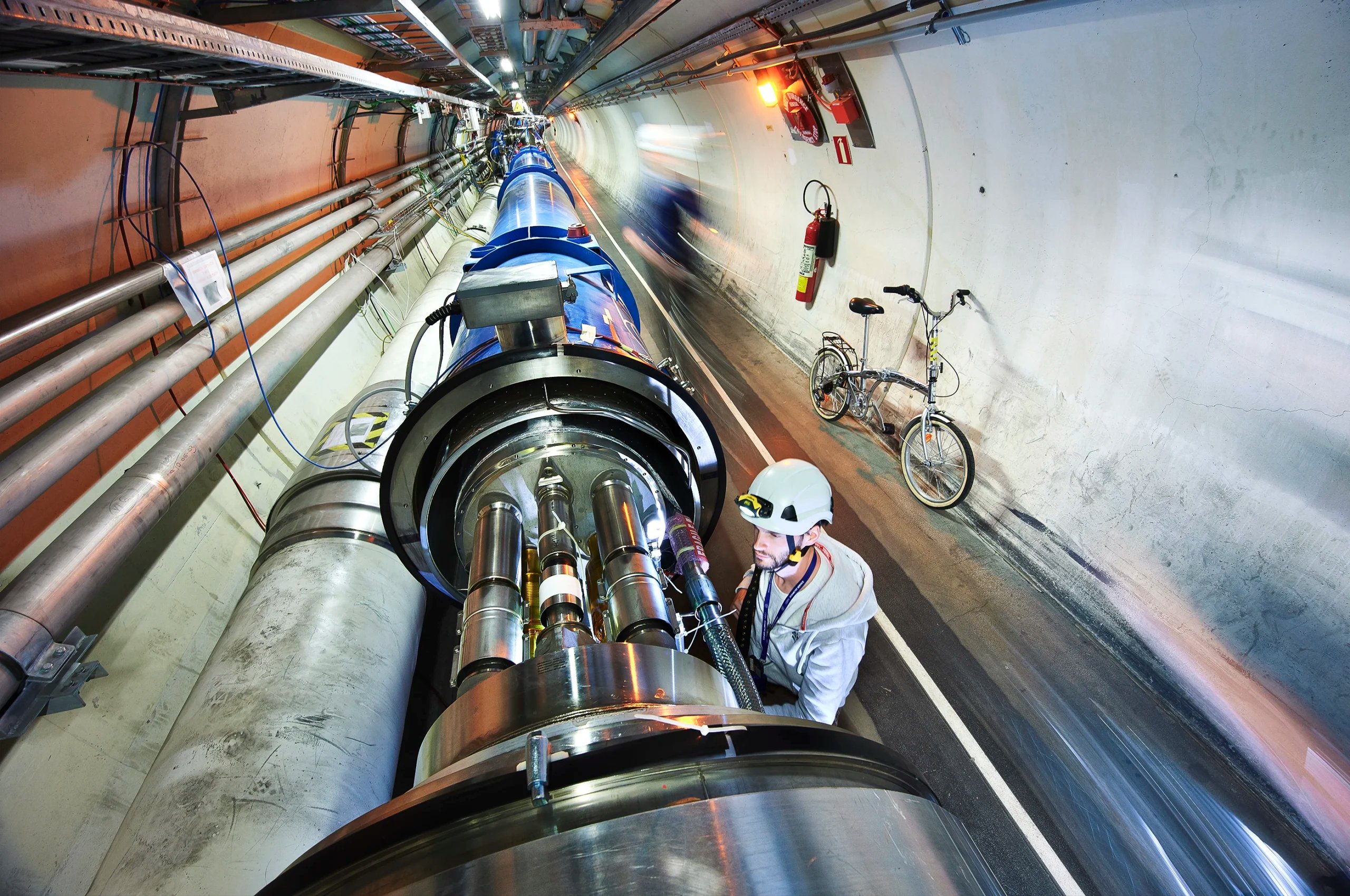 A long scientific tube in a tunnel is examined by a man in a helmet, a bike rests nearby.