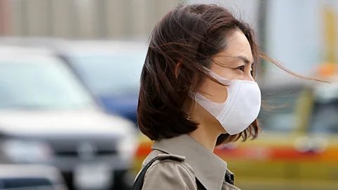 Getty Images A Japanese woman with short dark brown hair wears a white surgical mask. Cars are seen in the blurred background (Credit: Getty Images)