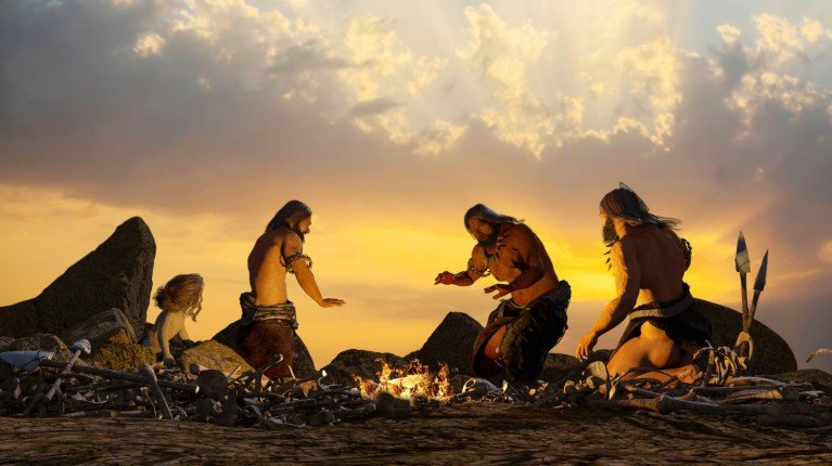 Realistic illustration of prehistoric people warming themselves by a campfire as the sun sets.