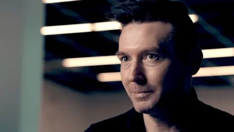 Matt Motyl, a man with brown hair styled in a quiff, pictured in close-up wearing a black shirt as he sits in an industrial-looking space with overhead strip lighting visible in the background blur.