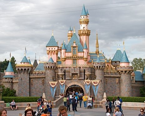 The pink and blue turreted castle of Disneyland, with tourists, under a cloudy sky.