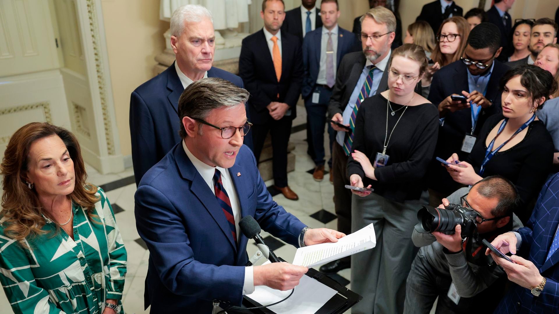 A man in a blue suit speaks at a lectern with papers as reporters and photographers crowd a government building hallway; a woman in a green patterned dress stands nearby.