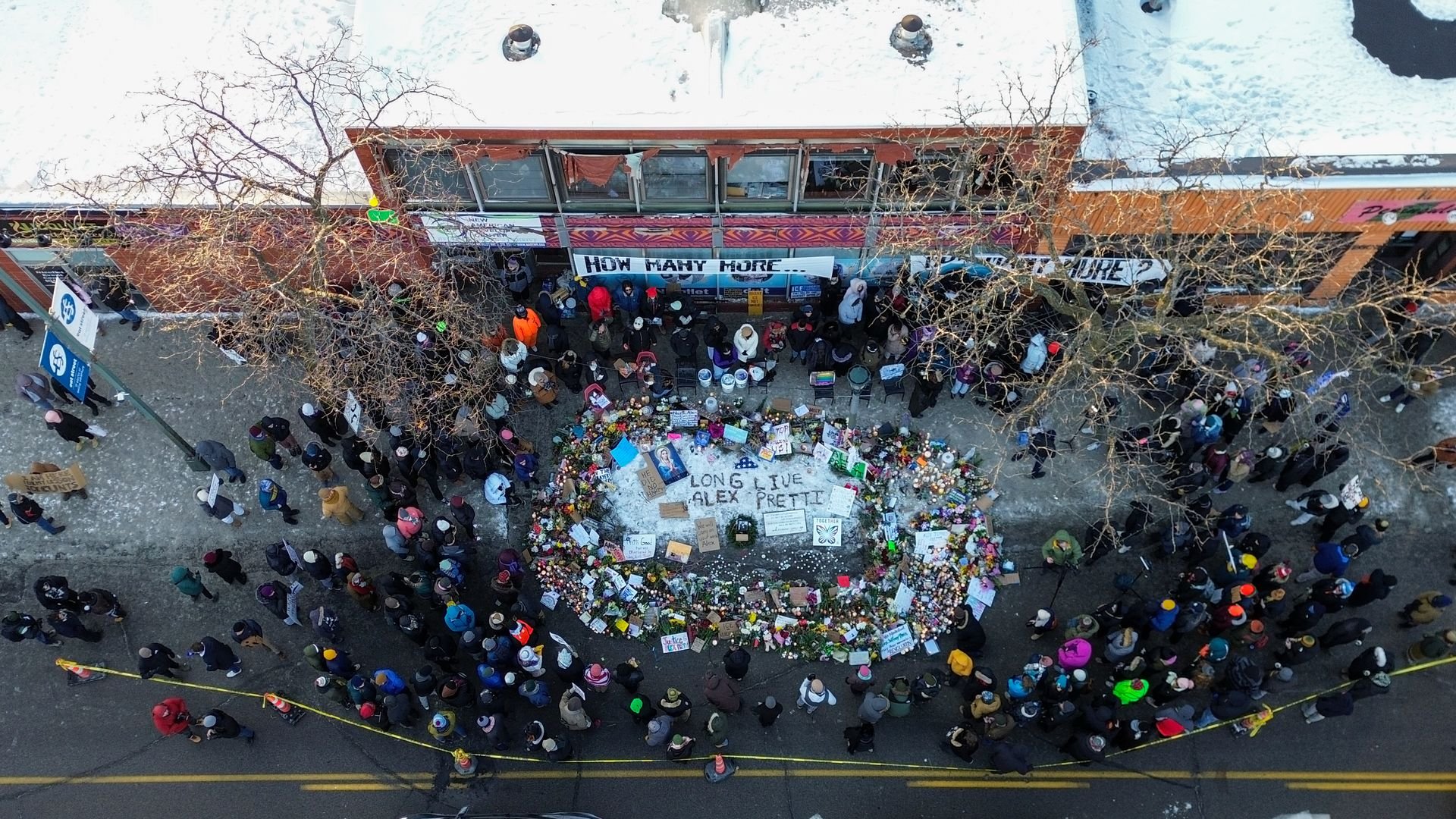 An overhead view of a group of people standing around a vigil for Alex Pretti, the man killed by a federal agent in Minnesota. Text on the ground reads 
