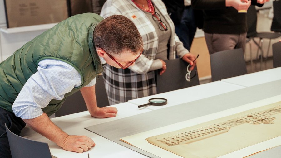 Person leans over a display case to closely examine an architectural drawing, with a magnifying glass placed nearby on the table.