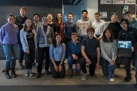17 people pose in two rows for a group photo. One holds up a laptop screen to include half-a-dozen remote participants.