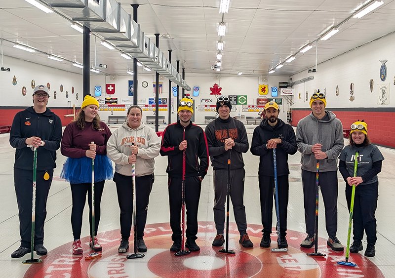 8 members of MIT's Curling team pose standing in a hockey rink.