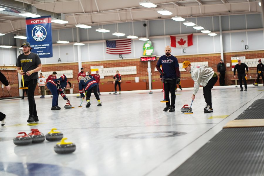 About a dozen men, some in uniform and some not, practice curling in a hockey rink