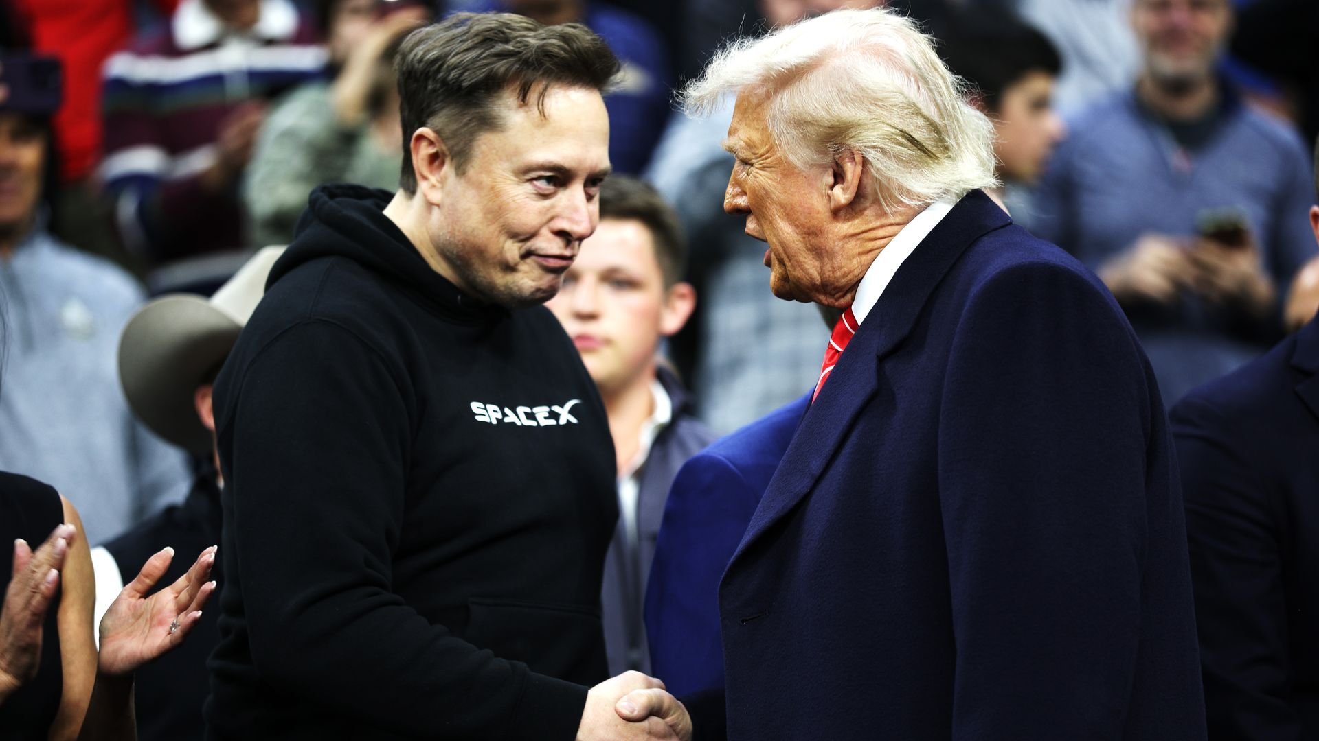 Elon Musk and Donald Trump, facing each other and shaking hands in front of a crowd.