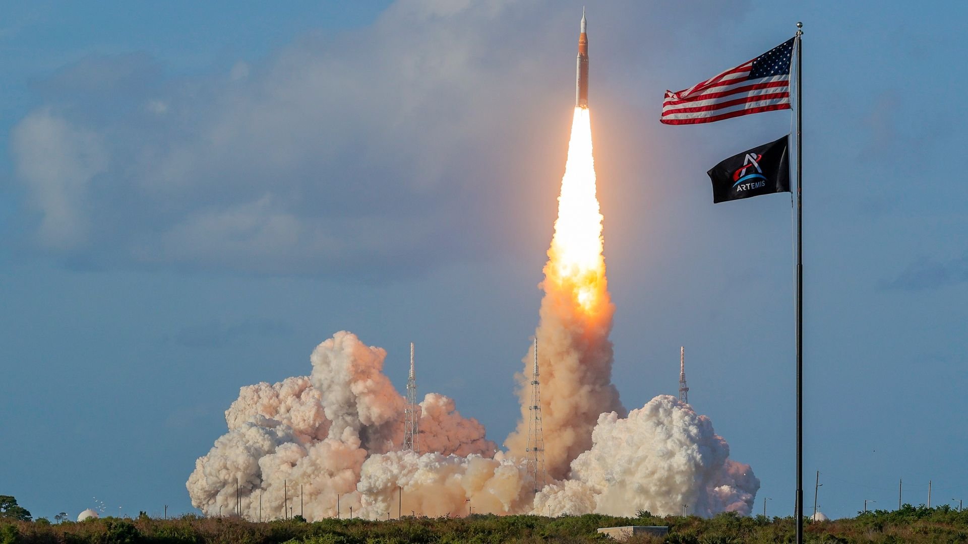 A rocket launches straight up with a bright flame and tall white smoke plume, against a blue sky. To the right, the American flag and an Artemis flag flap on a tall pole.