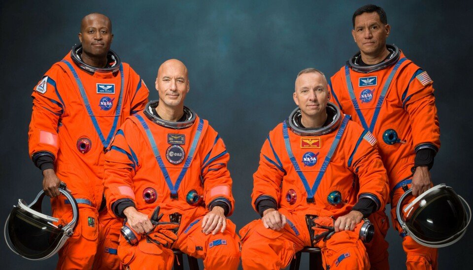 Four astronauts in orange spacesuits pose holding black helmets against a dark background.