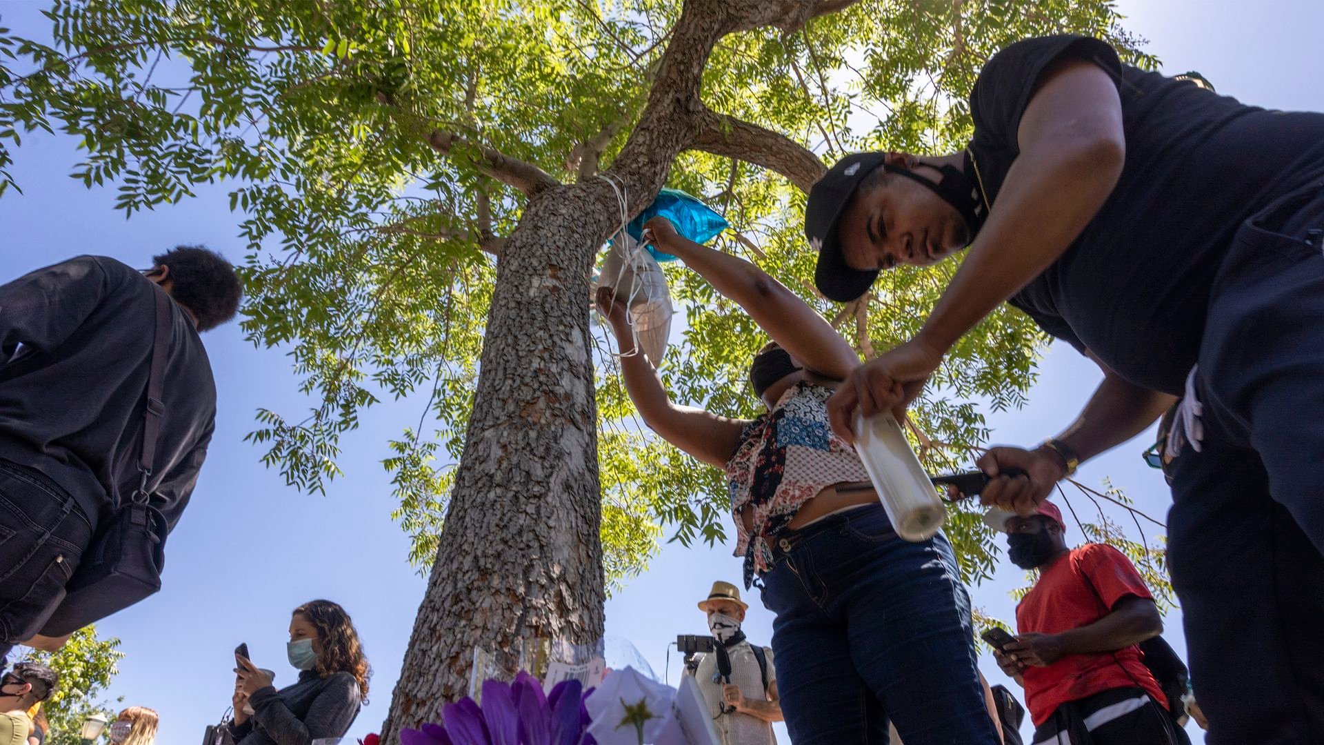 People wearing masks gather outdoors under a tree in daylight, hanging items on the tree and holding candles near colorful flowers, participating in a memorial or vigil.