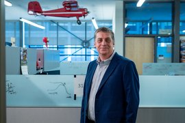 Jonathan How stands inside an office. A large red model airplane hangs from the ceiling behind him.