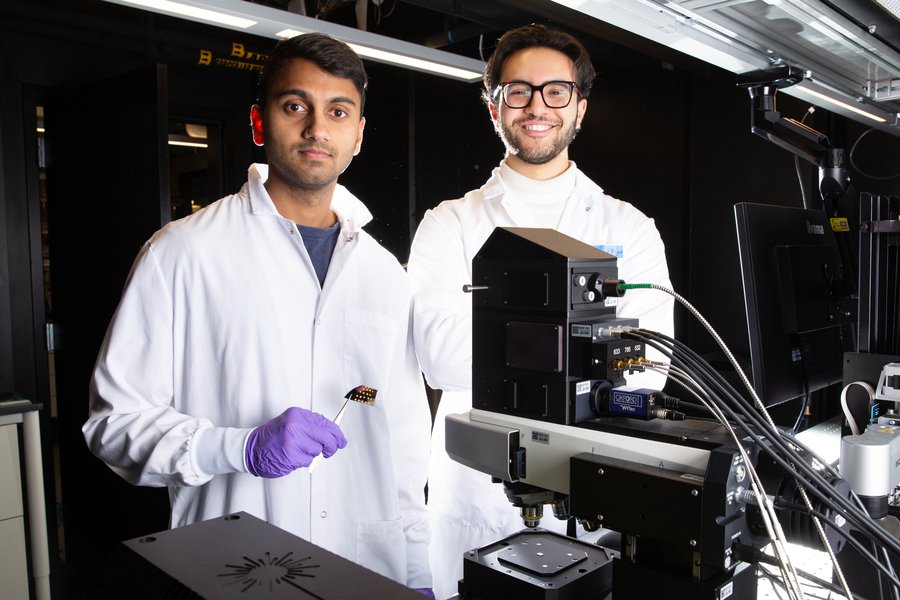 Aditya Garg and Seleem Badawy in lab with microscope. Garg holds a tiny device with tweezers.