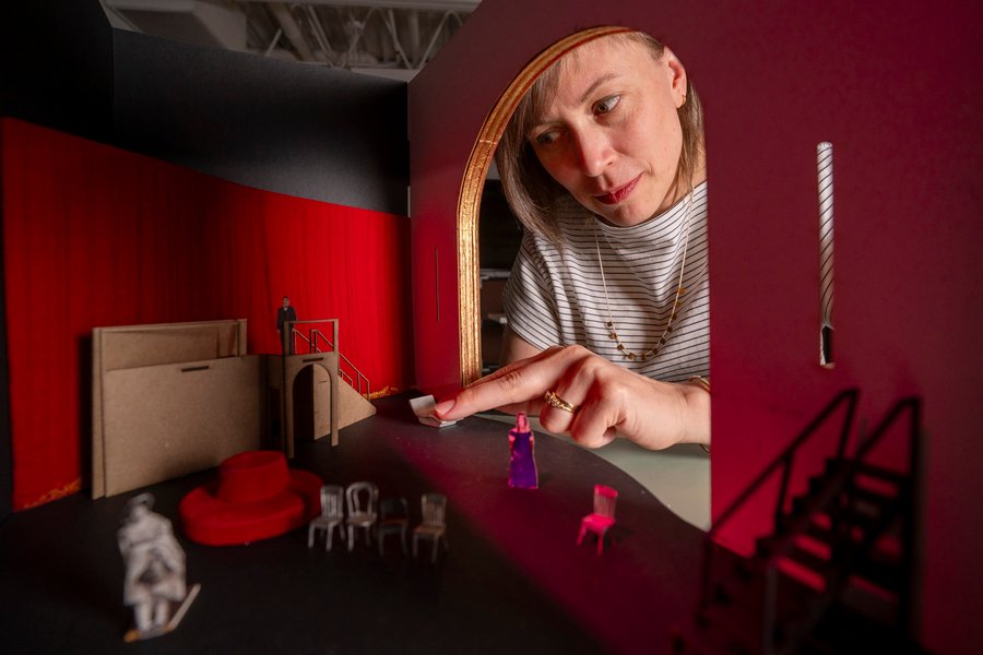 Sara Brown’s hand reaches through a tiny door into a miniature model of a red room with paper people and chairs.