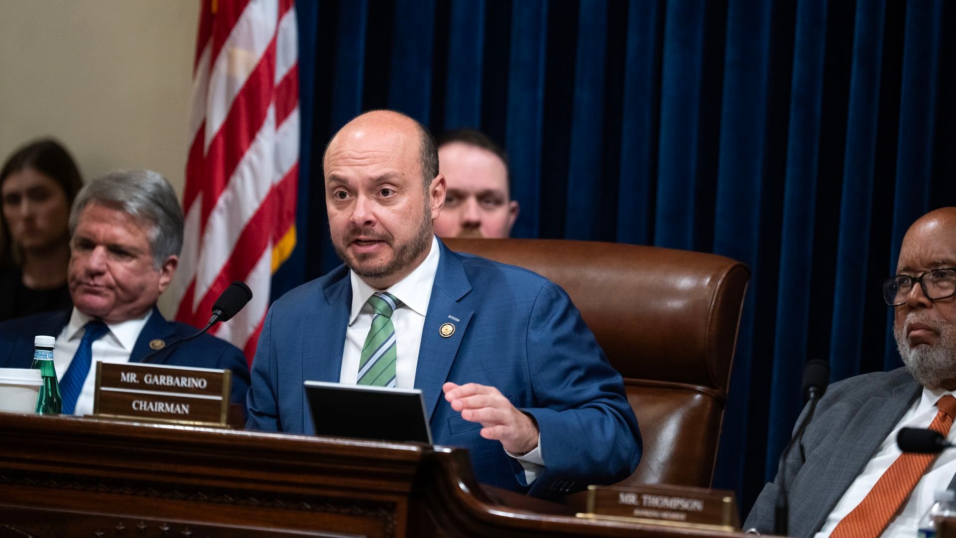 A man in a blue suit sitting in a chair behind a microphone with a nameplate that says 