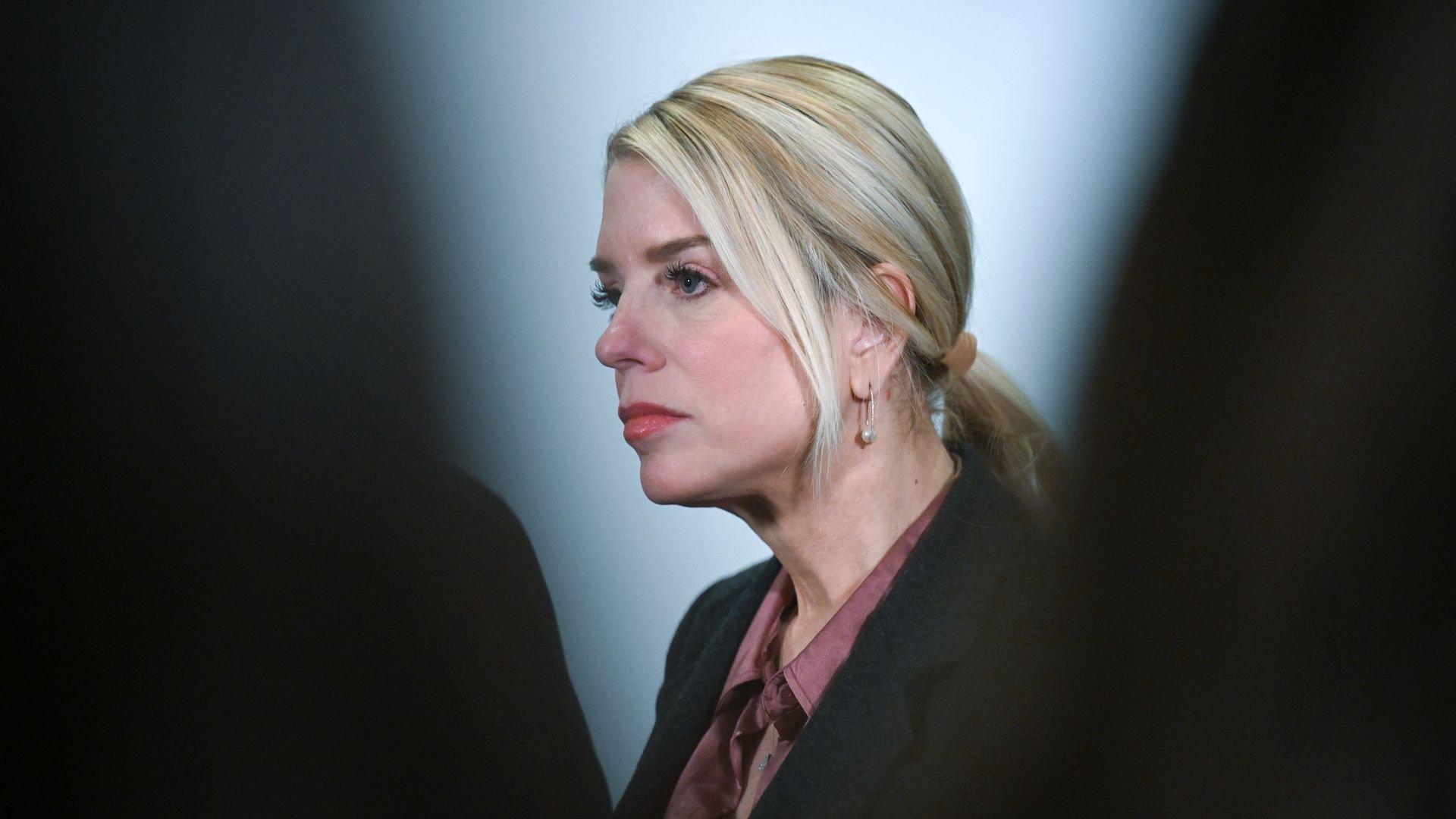 Pam Bondi, wearing her blonde hair in a ponytail and dressed in a dark jacket with a mauve collared blouse, looks on while answering questions from the media.