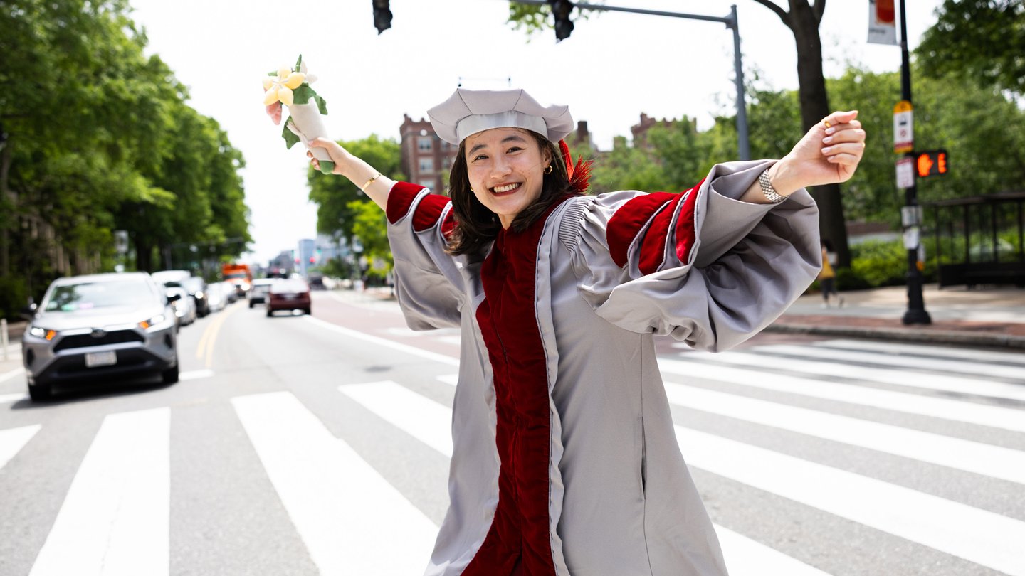 An MIT graduate in PhD graduation regalia smiles brightly as she crosses the street and holds a bouquet of flowers.