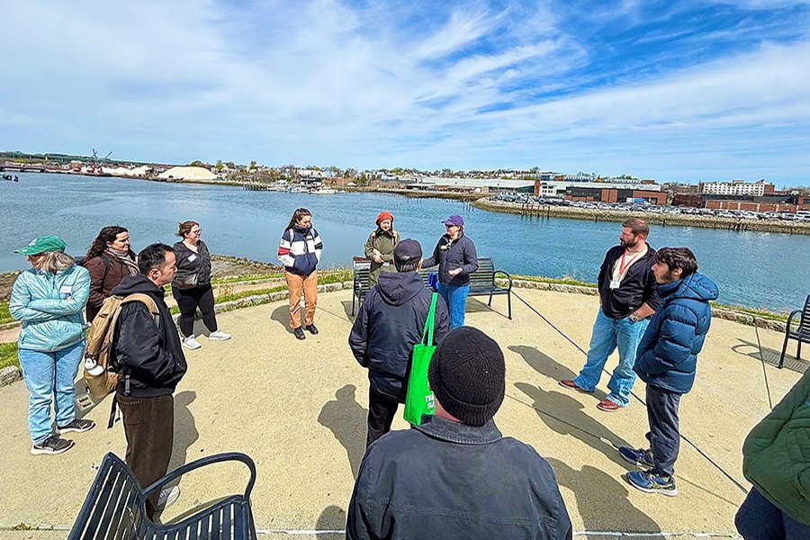 About a dozen people in winter outerwear stand in a circle on a seawall