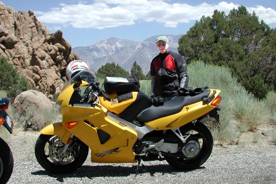Robert Liebeck stands next to a yellow motorcycle, posing on a road with a scenic backdrop.