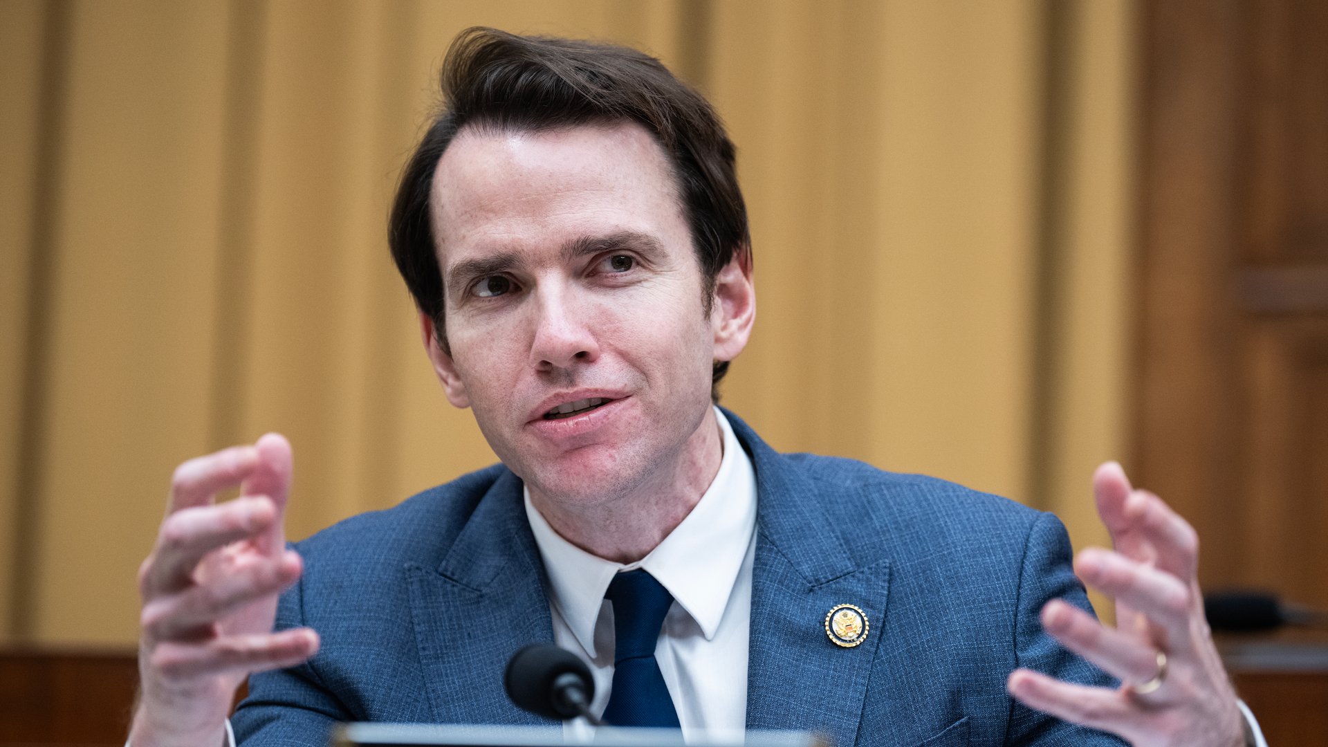 A man in a blue suit and dark tie gestures with hands while speaking at a formal setting, seated behind a nameplate labeled 