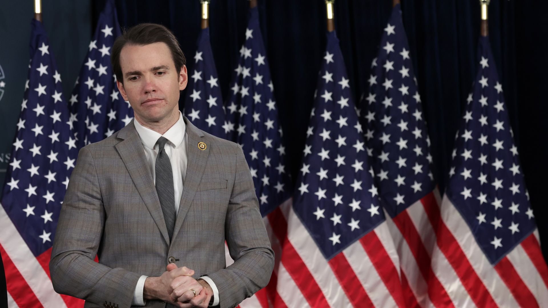 A man in a gray suit and tie stands in front of six American flags with gold eagle finials against a dark blue background, looking thoughtful with hands clasped.