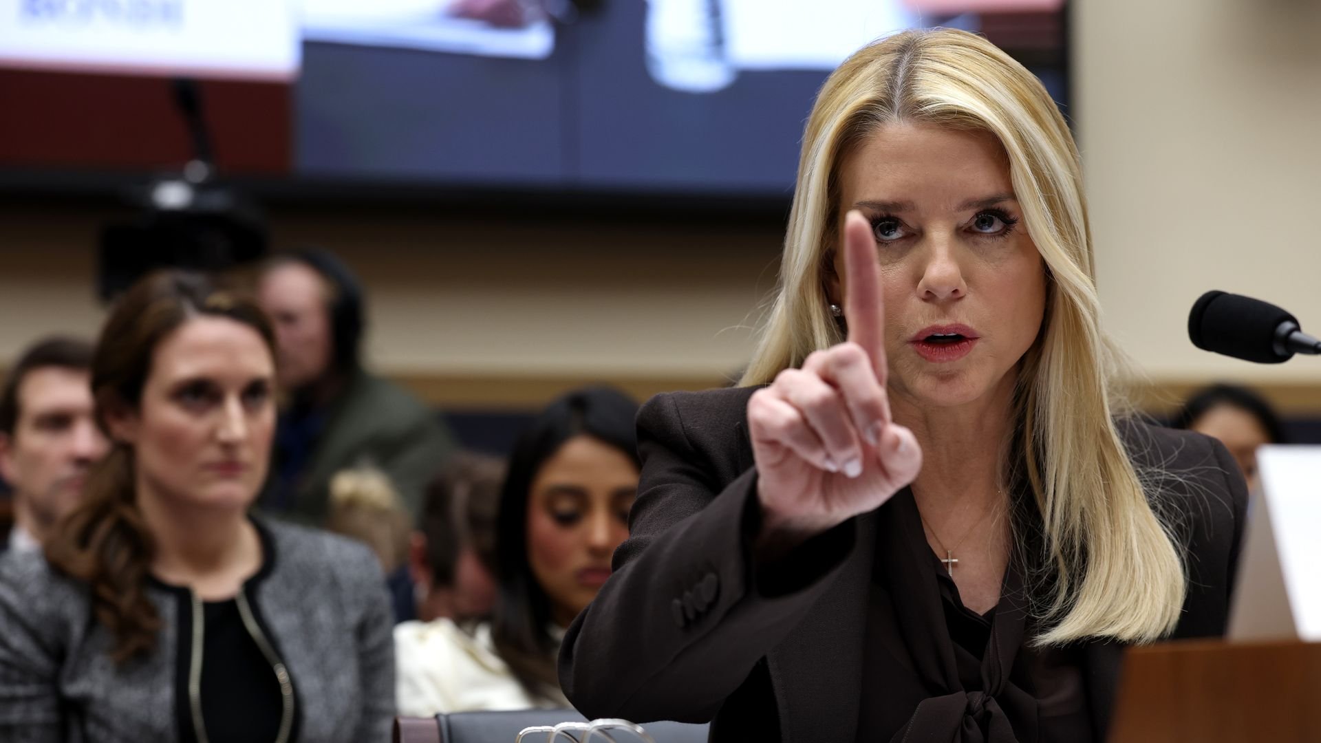 A blonde woman in a black jacket gestures with her index finger raised while speaking at a hearing, with a microphone and large binders on a table in front of her; blurred audience in the background.