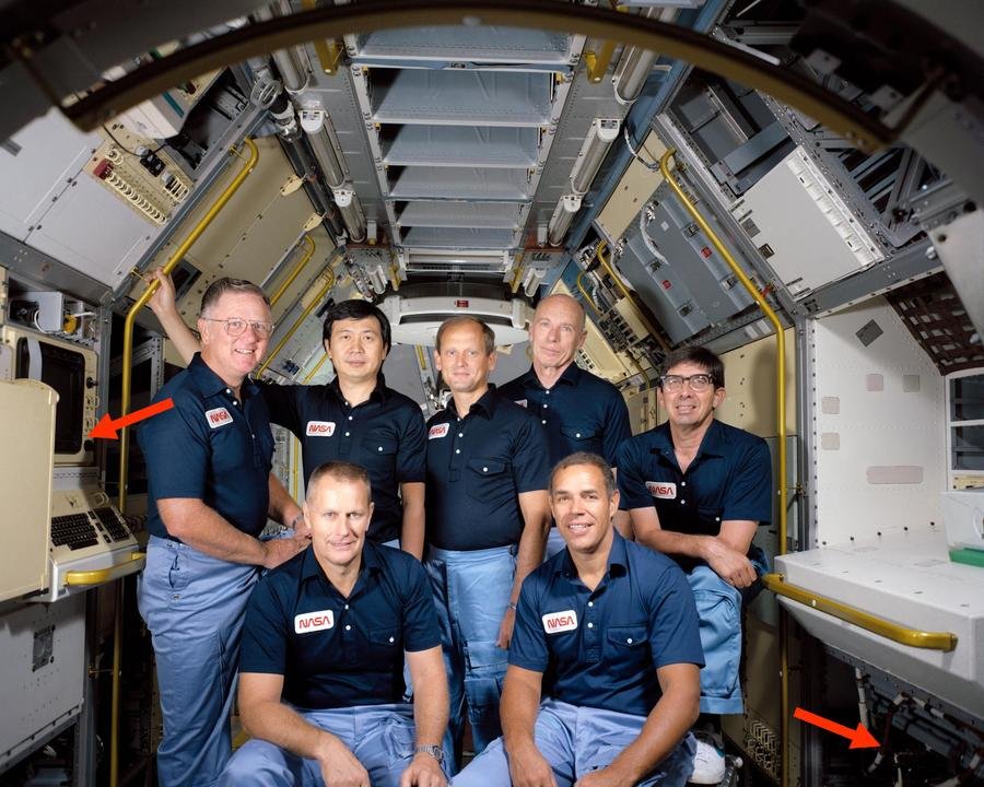 This photo shows astronauts inside Spacelab (but not in space). The Spacelab computers were mounted under the Work Bench (right arrow). The Data Display System (left arrow) provided the interface to the computers. Photo is STS-51B Crew Portrait, 1984.