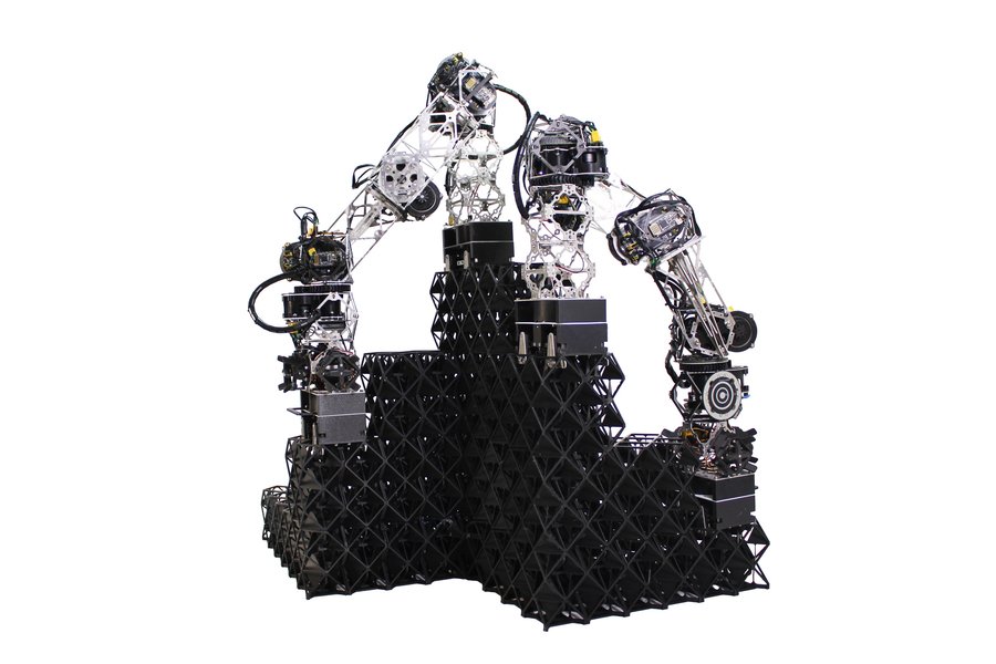 Two robot arms stand on top of a voxel structure as they continue to build it up.