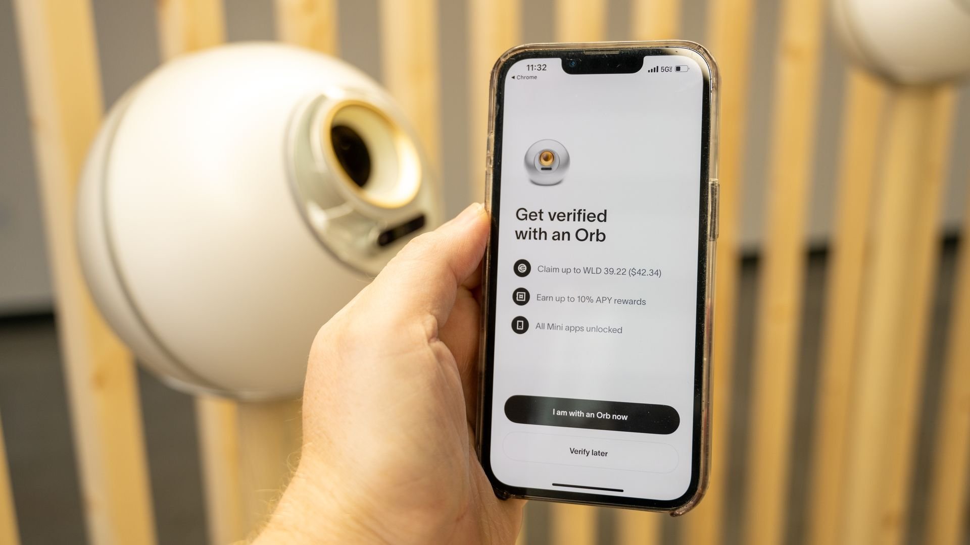 Image of a hand holding a smartphone displaying World app for Worldcoin identity verification with orb scanning device in background.