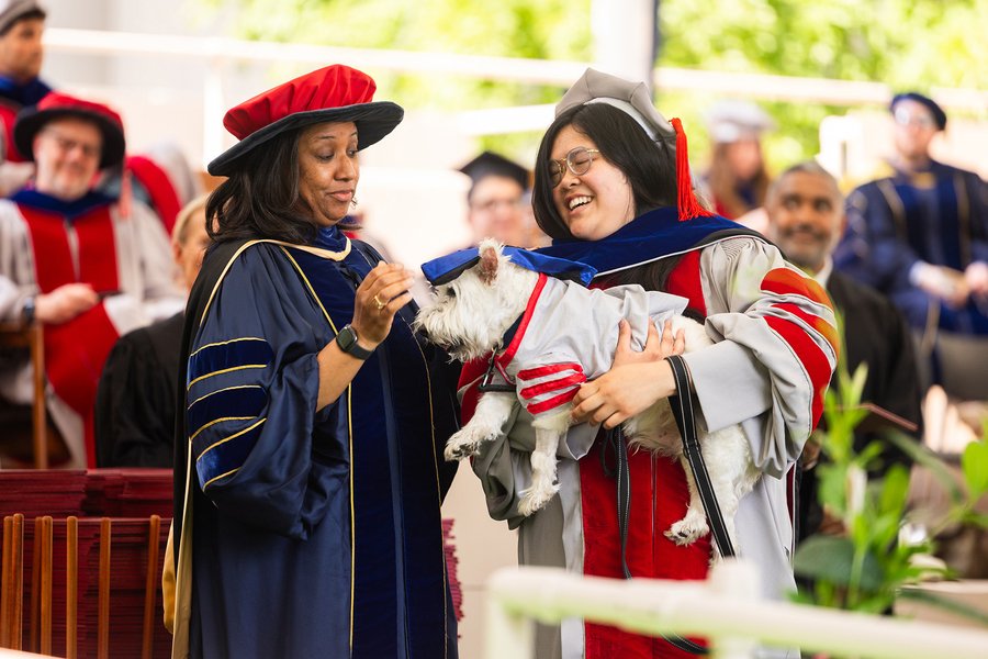 Michelle Quien holds her dog, a white terrier, while both are dressed in graduation regalia and face Kristala Prather.