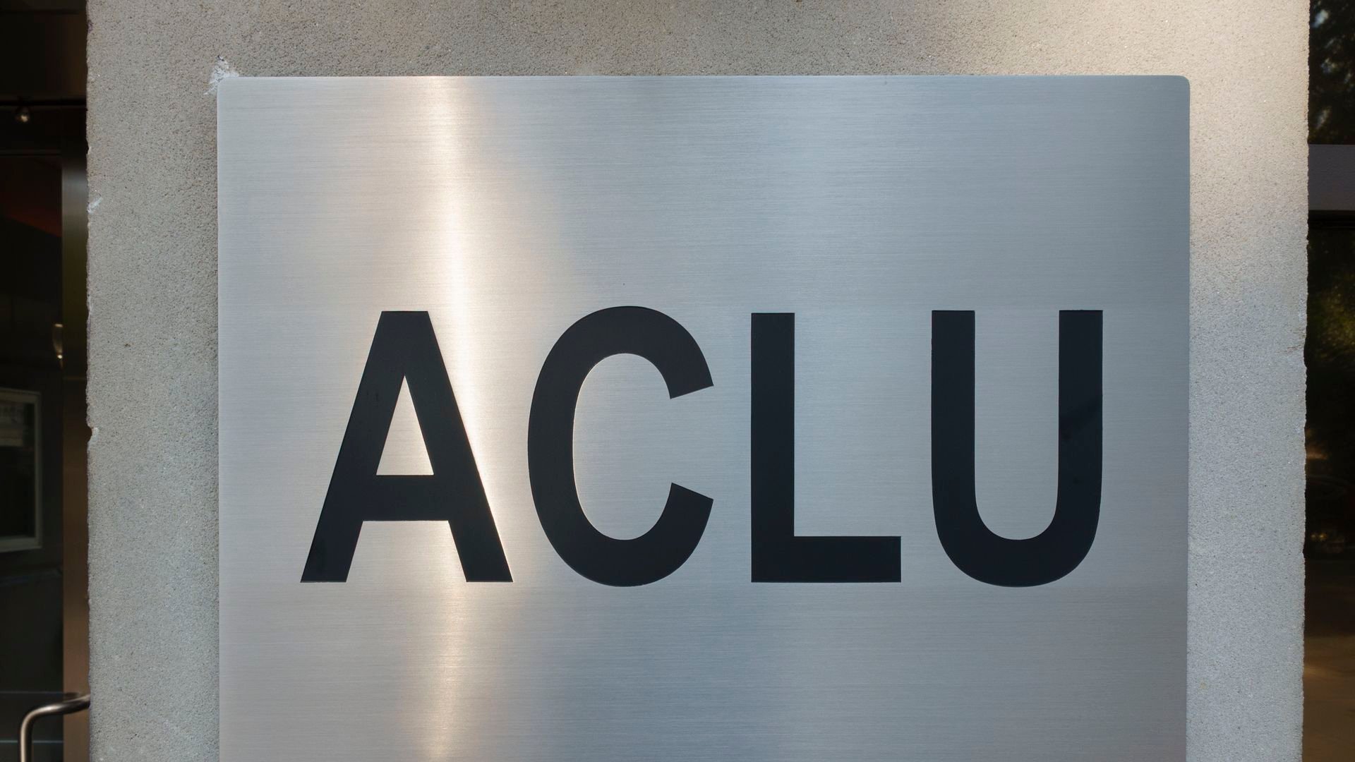 Silver metallic sign with large black letters spelling ACLU mounted on a beige wall outside a building entrance.