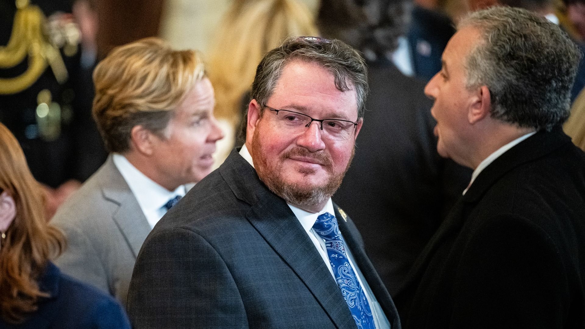 Group of people in formal attire attending an event, featuring a man with glasses and a blue paisley tie looking to the side amidst conversations in a crowded indoor setting.