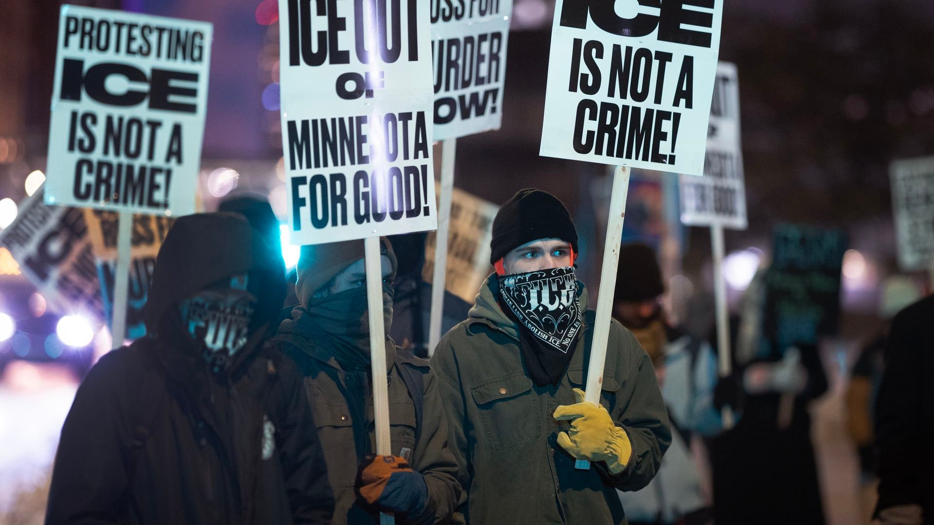 Protesters wearing winter clothes and face coverings hold signs at night saying 