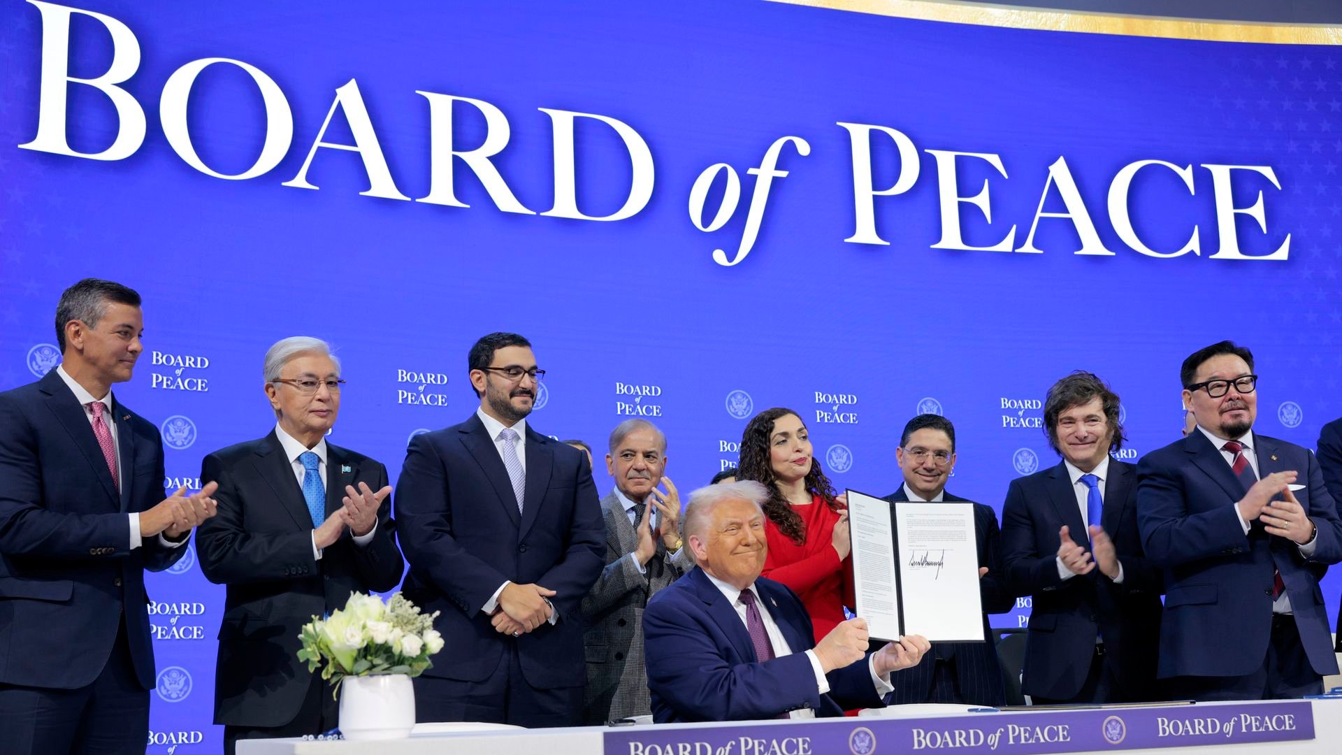 Blue Board of Peace background with line of people in front of it and man sitting at table in front of them
