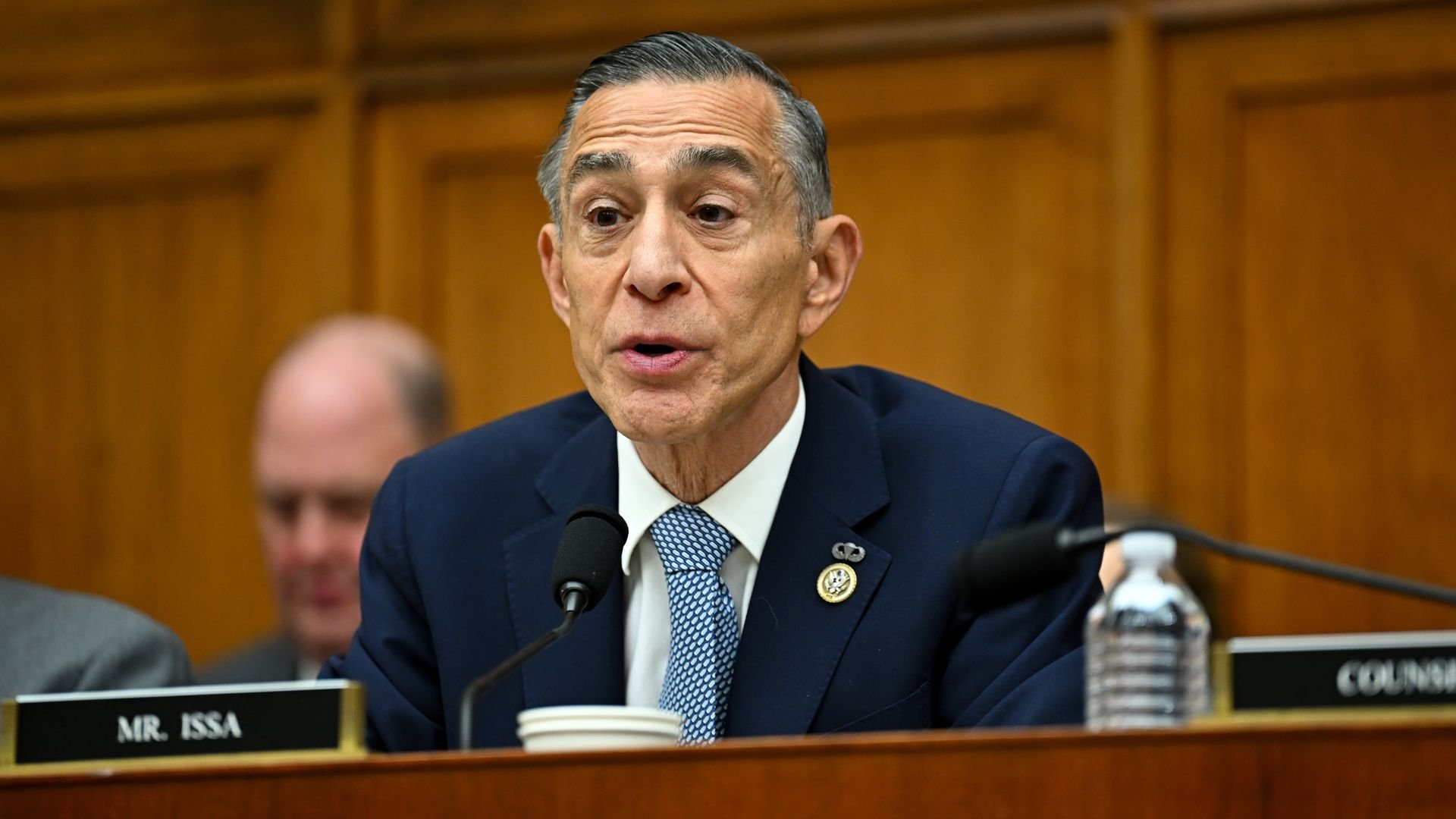 Man in a navy blue suit and patterned blue tie speaking into a microphone during a formal meeting with wood-paneled walls and a nameplate labeled 
