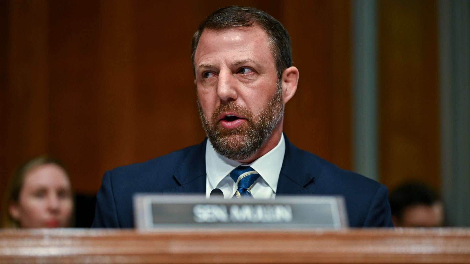 A bearded man in a dark blue suit and striped tie, Senator Markwayne Mullin, speaks at a wooden panel desk in a formal hearing, with a blurred audience behind; a nameplate reads 