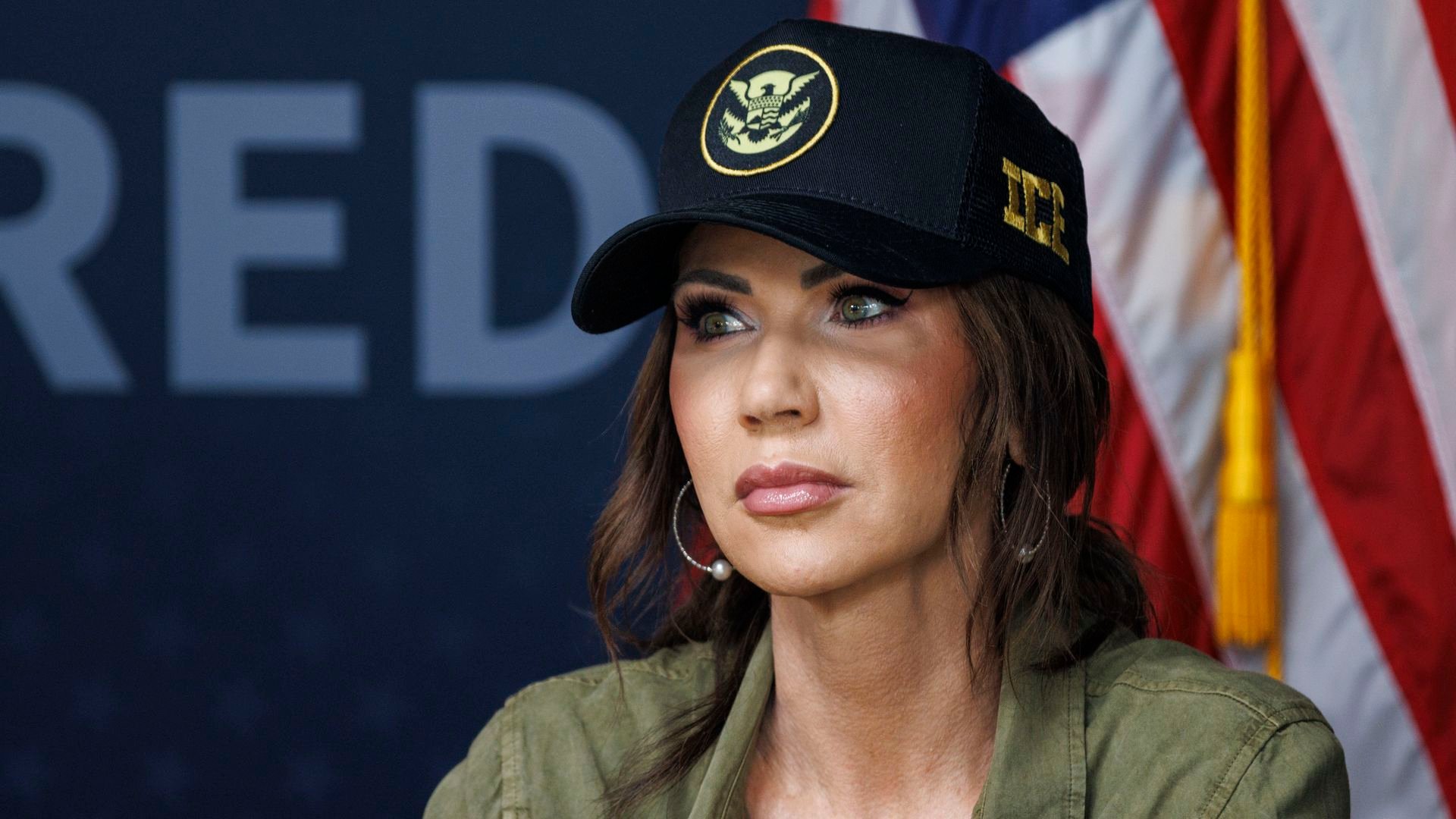 Woman with green eyes wearing a black hat with a gold emblem and an olive green shirt, in front of an American flag and a dark background with partial white text.